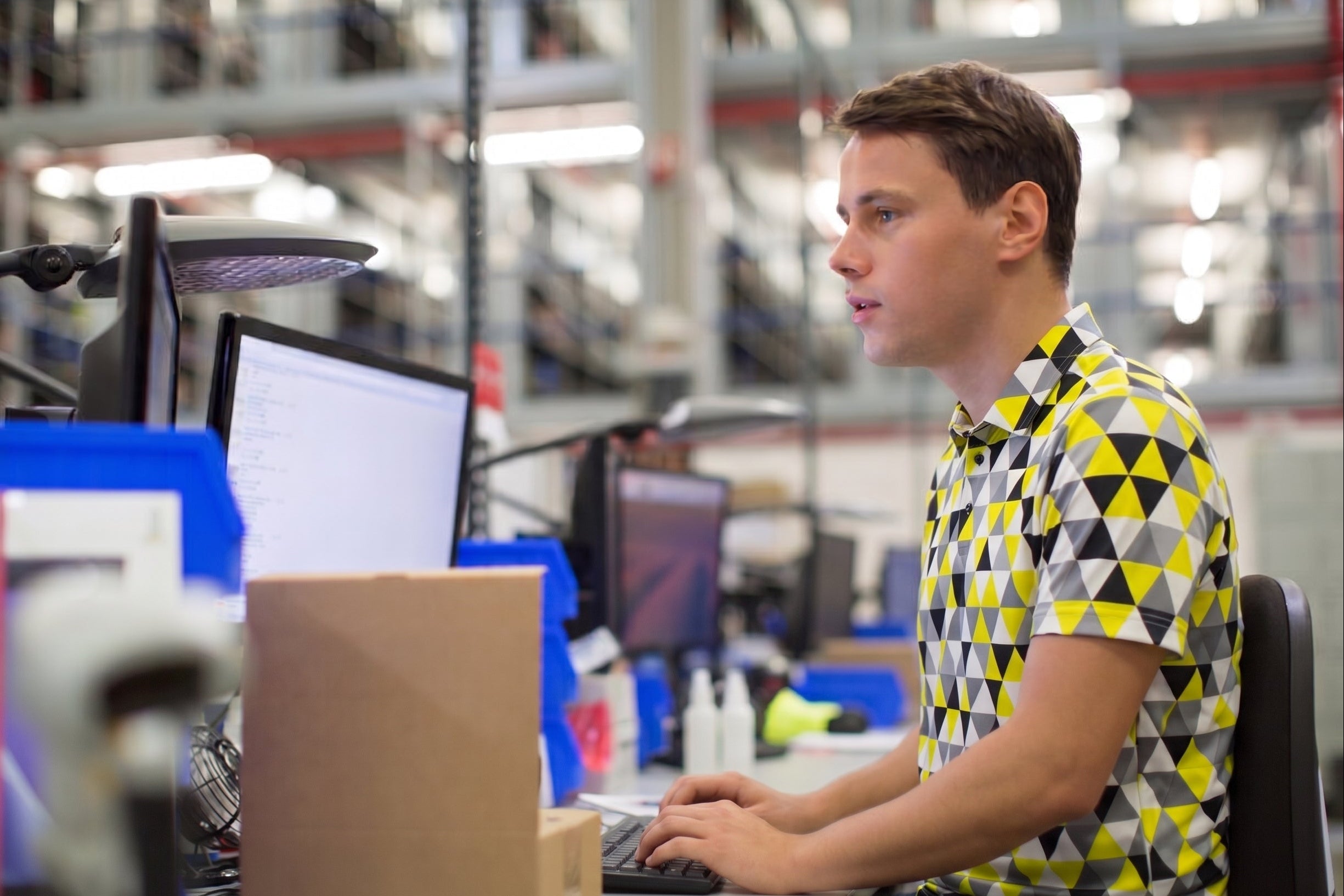 A man in a yellow, black, and white geometric patterned short-sleeve shirt sits at a desk in a factory or workshop environment, looking at a computer monitor.